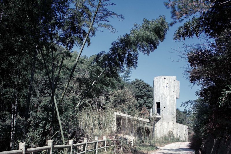 Board-formed concrete tower rising above a single-story volume along a gravel path framed by trees