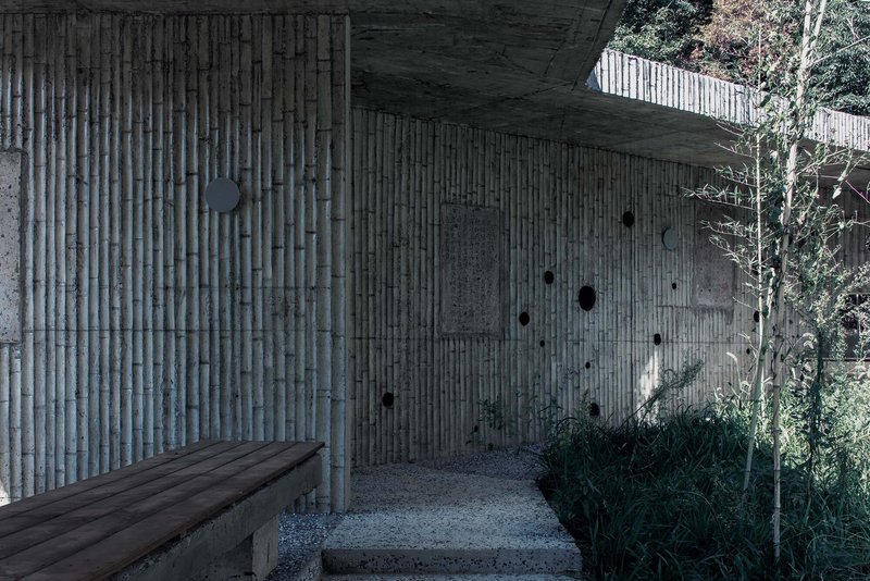 Covered entry courtyard with vertical timber formwork walls and gravel floor surrounded by planted vegetation