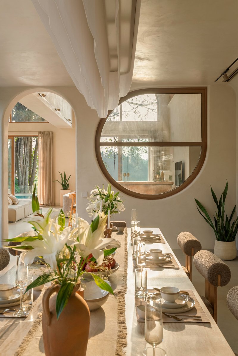 Dining table beneath a large circular window framing trees, with sculptural ceiling plane above