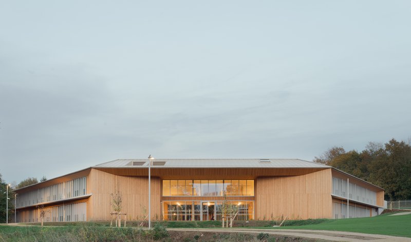 Timber-clad building with folded roof and glazed entry set against lawn and autumn treeline