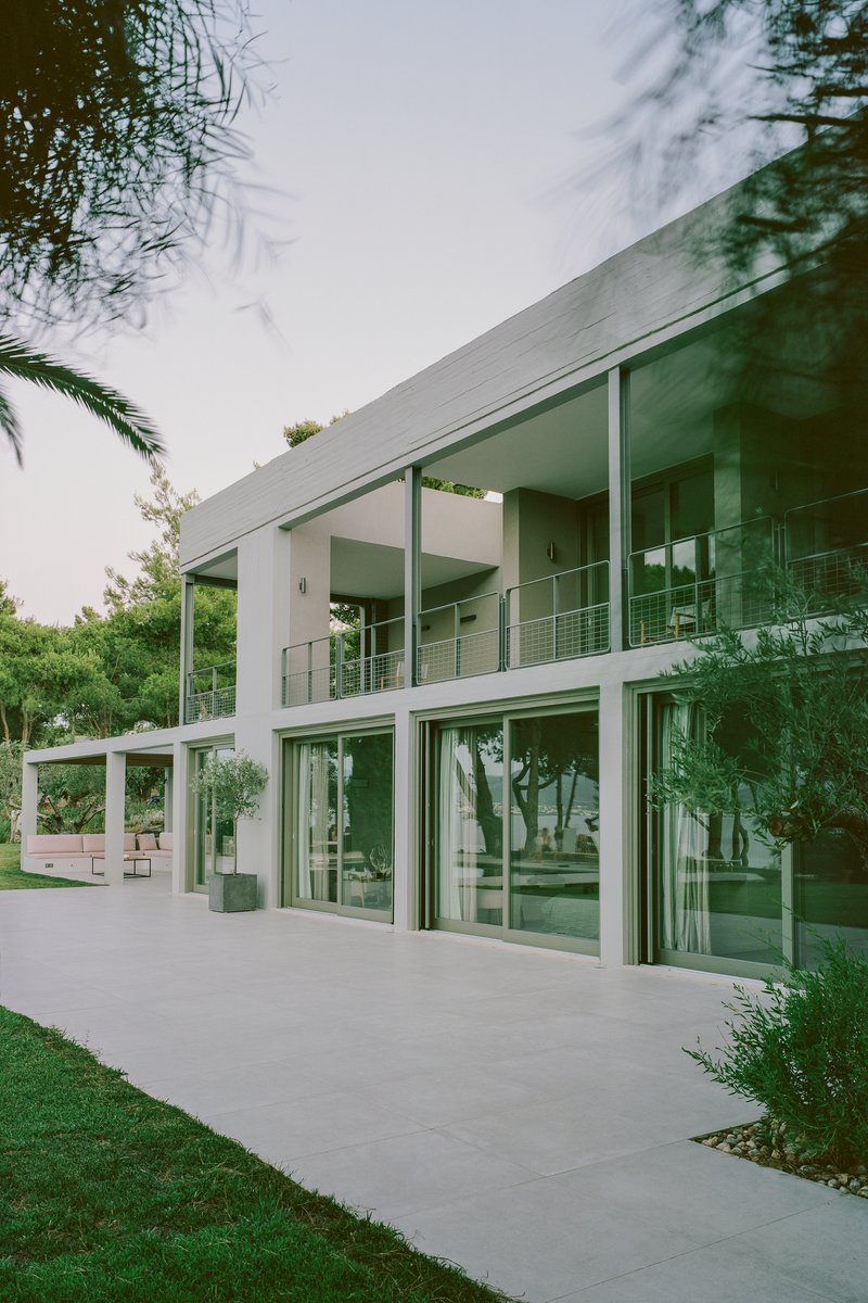 Two-story glass facade with white structural columns framed by lawn and palm fronds overhead