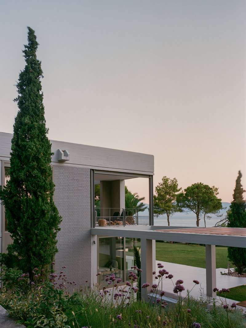 Corner elevation showing grey brick volumes and cantilevered terrace with cypress trees and purple flowering plants