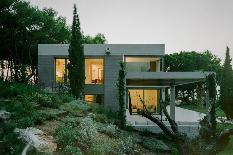 Evening view of illuminated grey brick volumes set into sloped terrain with native shrubs and trees