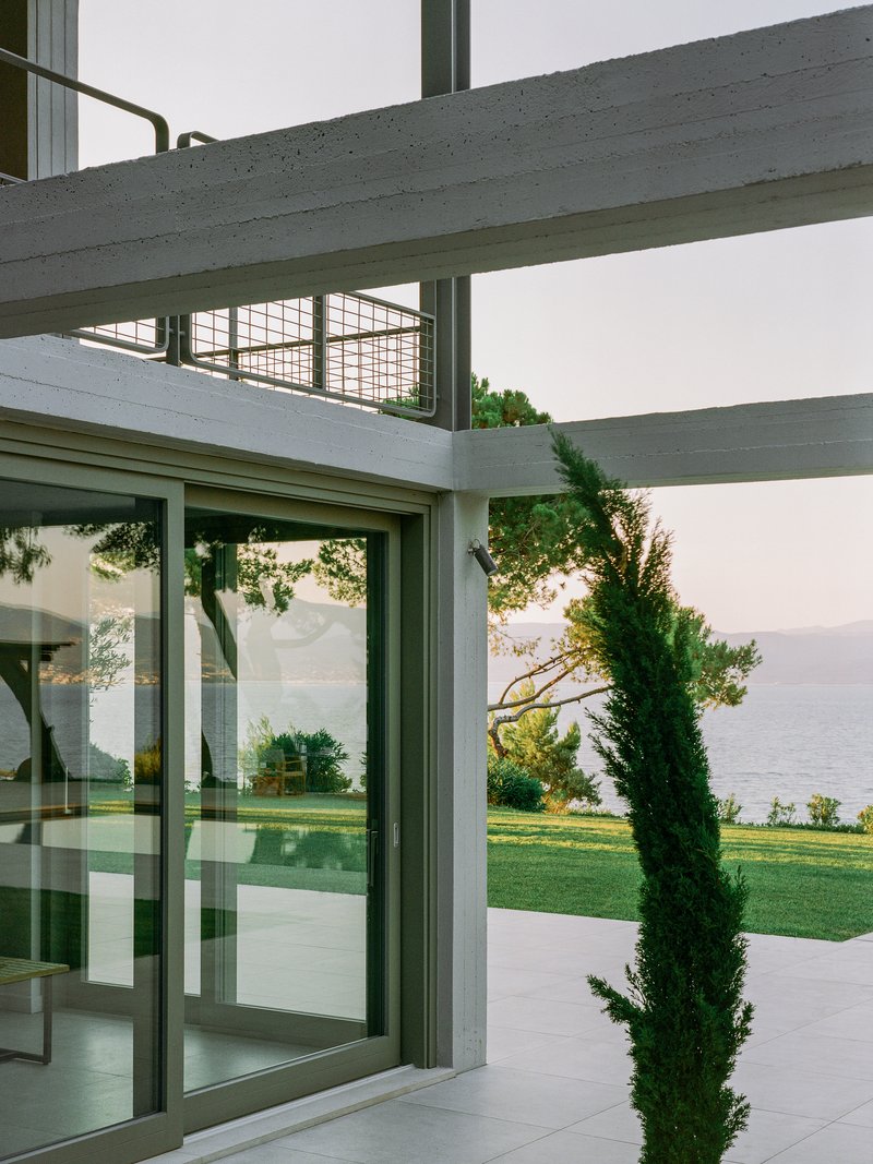 Concrete beams and glazed sliding doors opening to lawn with waterfront and cypress beyond