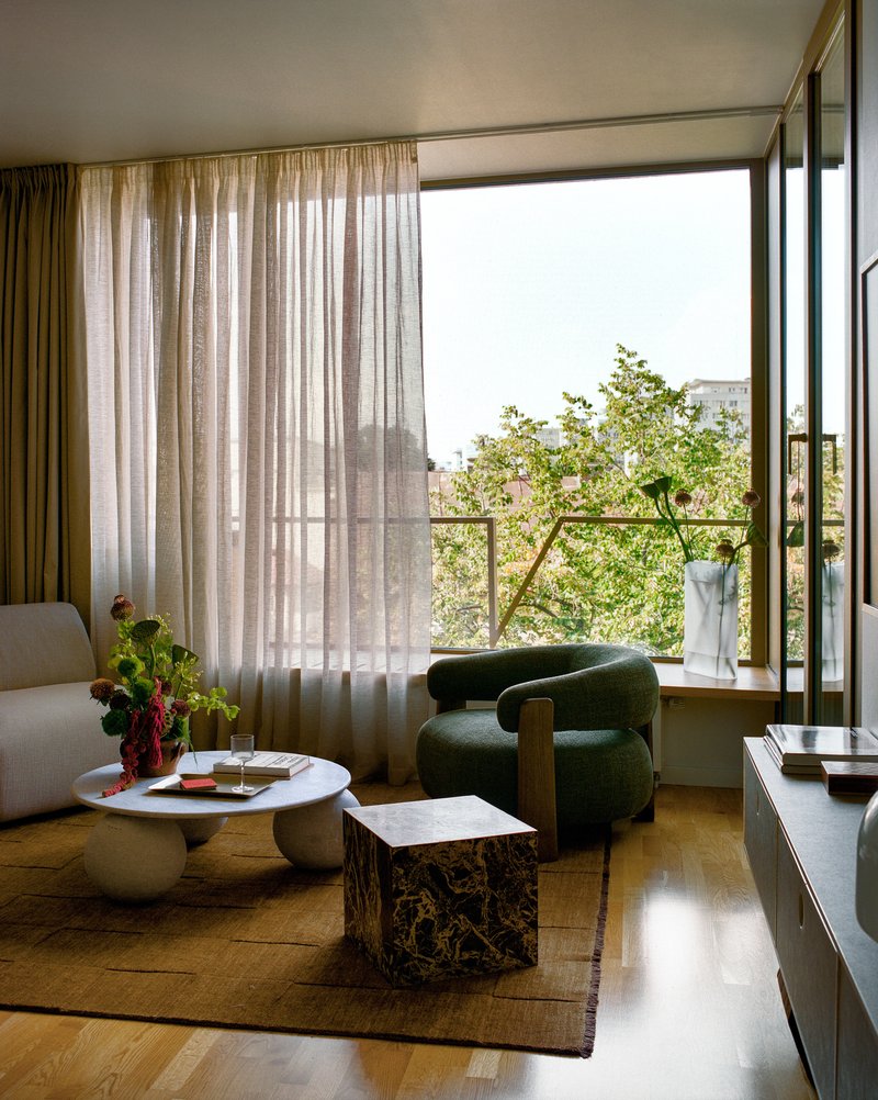 Living room with sheer curtains and open balcony door overlooking treetops and distant buildings