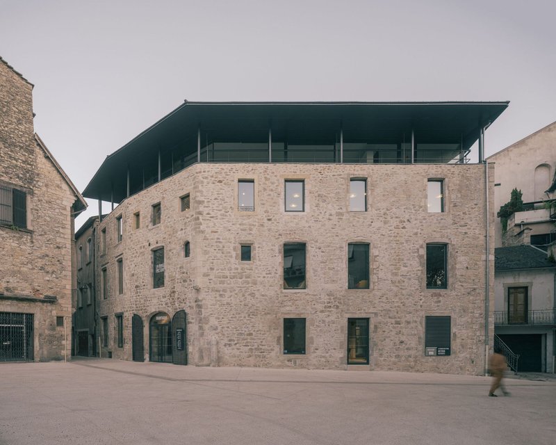 Stone facade with irregularly placed windows and a floating dark roof canopy under overcast skies