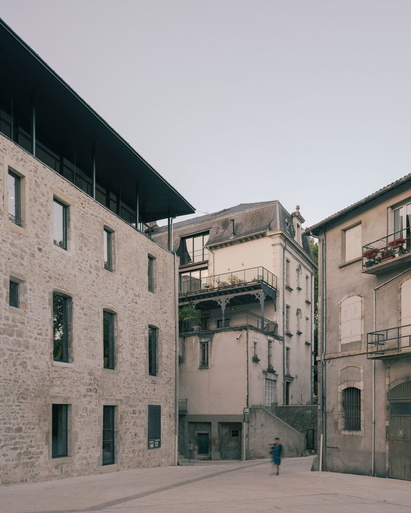 Courtyard view showing restored stone facade with a glazed upper volume topped by a dark metal canopy