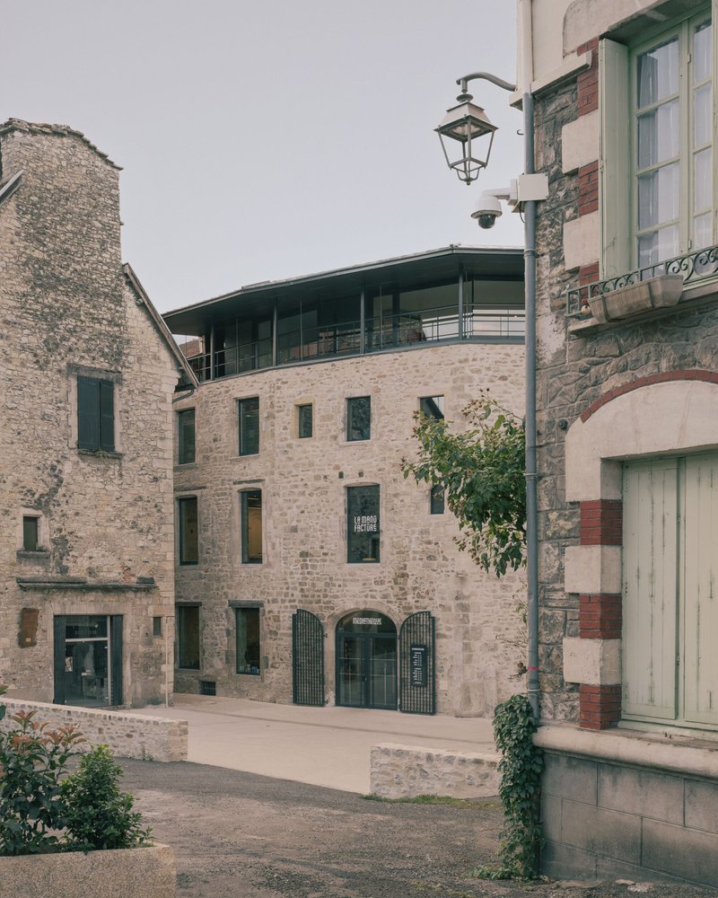 Narrow plaza framed by stone buildings with arched openings and a small tree between paving stones