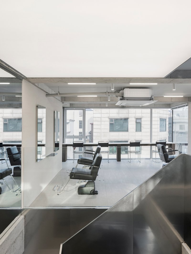 Salon floor with rows of barber chairs facing full-height glazing and exposed concrete ceiling