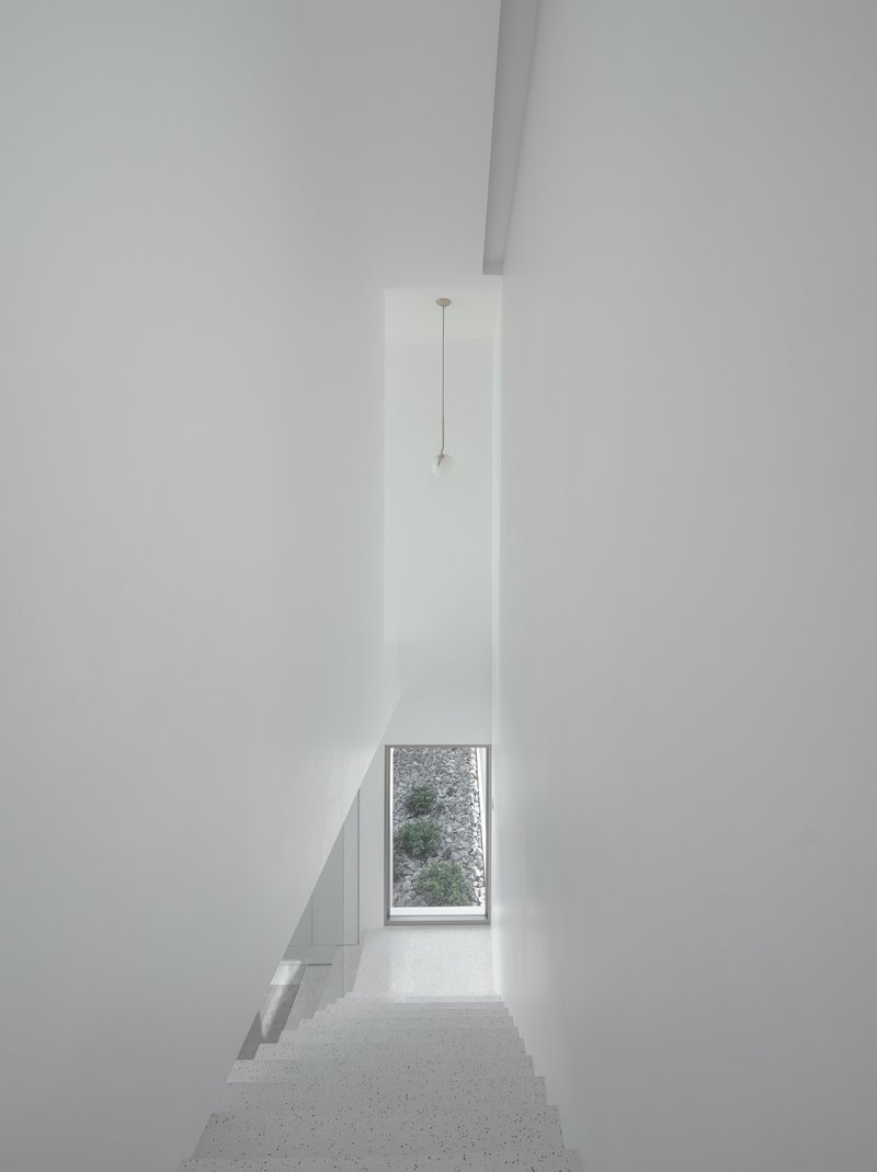 Narrow white stairwell with terrazzo treads descending toward a tall window framing greenery