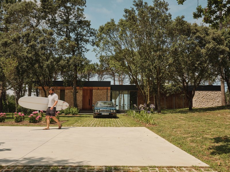 Street view of the single-story residence with timber and stone volumes set among mature trees