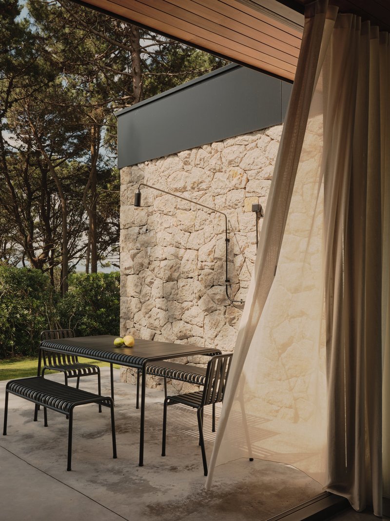 Outdoor terrace with table and benches facing a coursed stone wall with wall-mounted shower under timber soffit