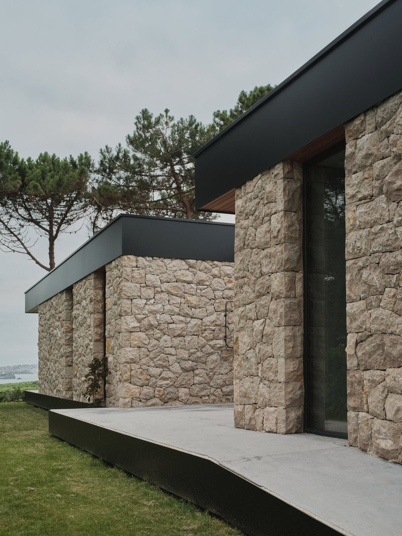 Exterior view showing the stone wall volumes capped with dark metal fascia beside a coastal lawn and pine tree