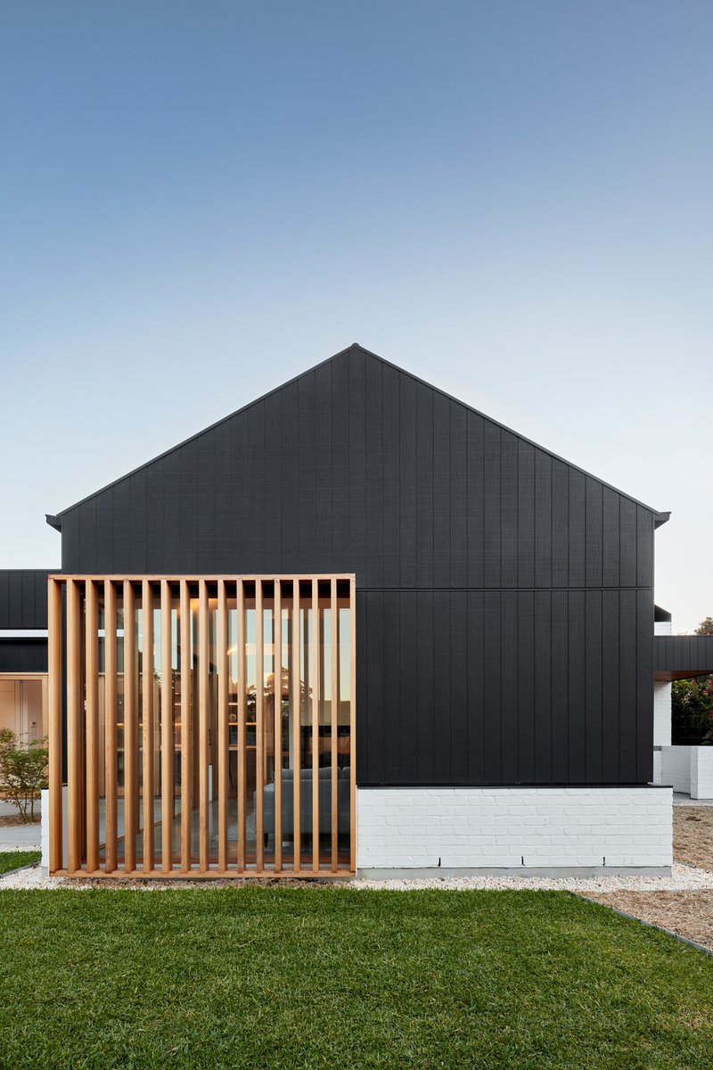 Gabled black timber-clad volume with vertical timber screen over white brick base and lawn