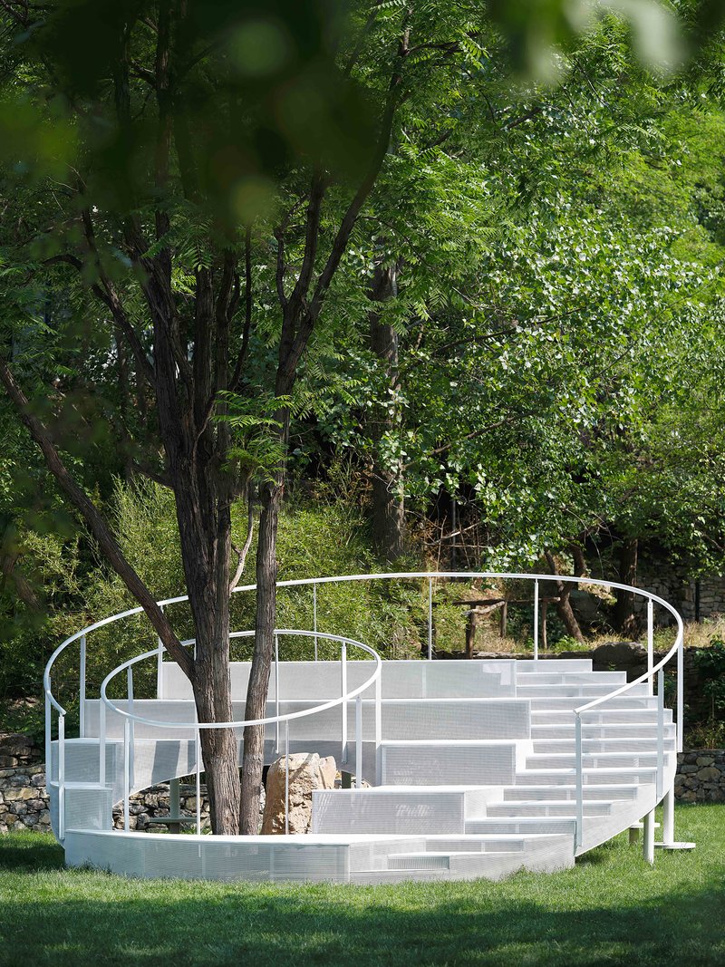 White steel amphitheater structure with tiered seating wrapped around existing trees in a summer lawn