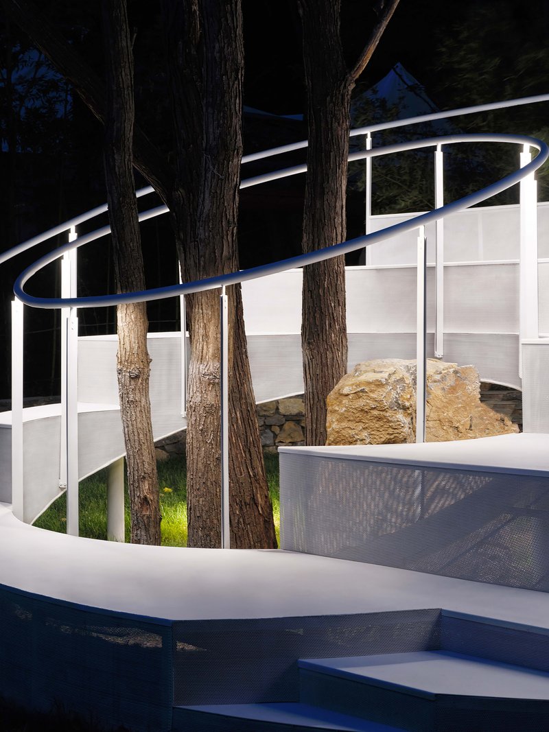 Night view of curved white stairs and perforated metal balustrade weaving between tree trunks