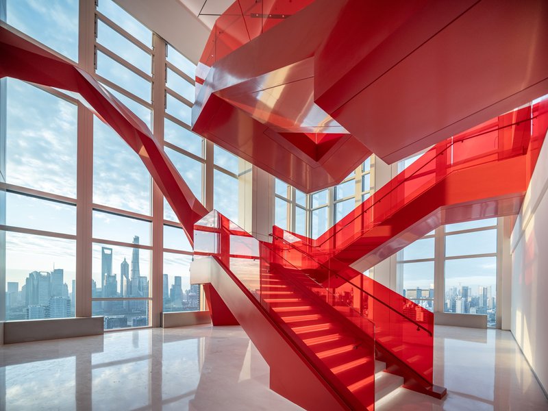 Double-height interior staircase wrapped in translucent red glass with floor-to-ceiling curtain wall beyond