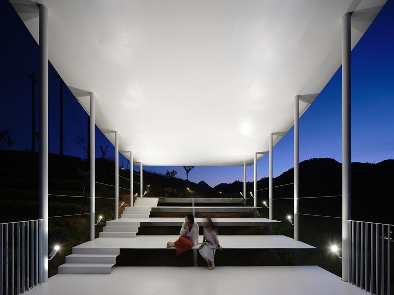 Tiered amphitheater seating under an illuminated canopy with columns at night as two people sit together