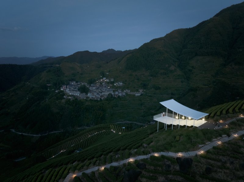 Aerial view of the pavilion perched on terraced tea fields above a hillside village at twilight