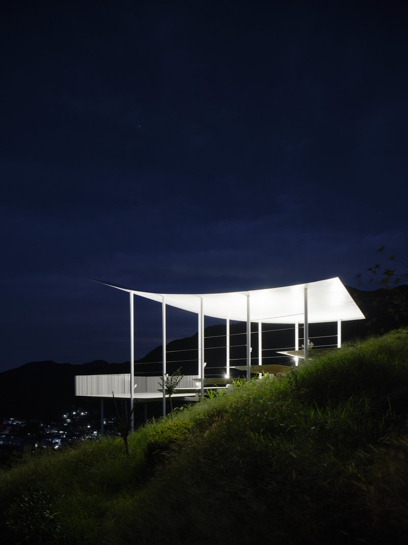 Side view of the illuminated pavilion with curved roof on a grassy slope at night