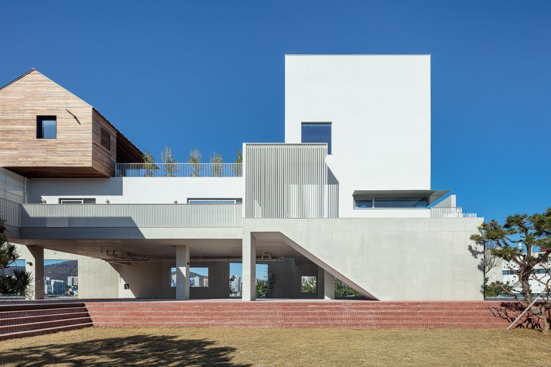 Elevated concrete structure with stacked volumes in timber, white corrugated metal, and stucco above a red brick platform and lawn