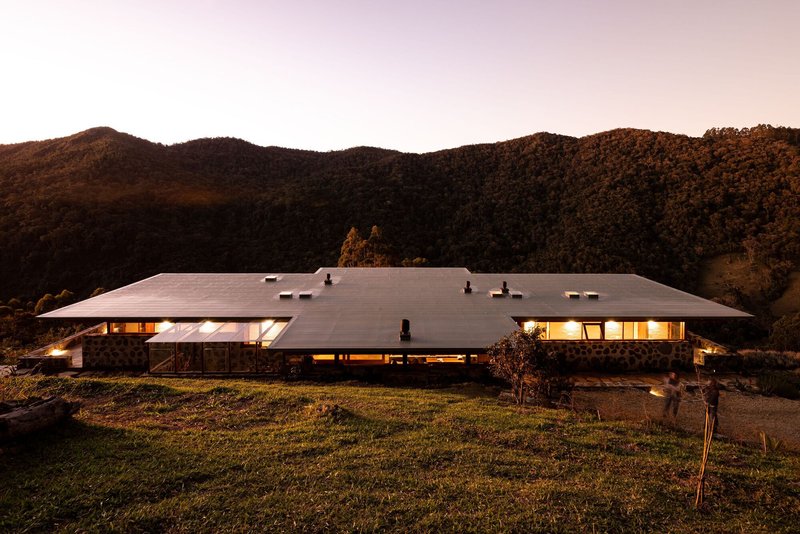 Low-slung residence with broad overhanging roof set against forested hillside at dusk