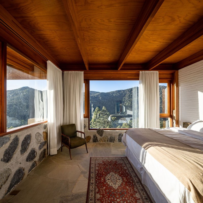 Bedroom with corner windows framing mountain views and river stone half-wall beneath timber-clad ceiling