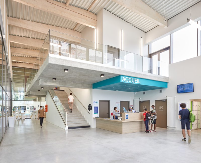 Double-height reception lobby with timber-slatted ceiling, mezzanine walkway, and visitors at the wooden desk below