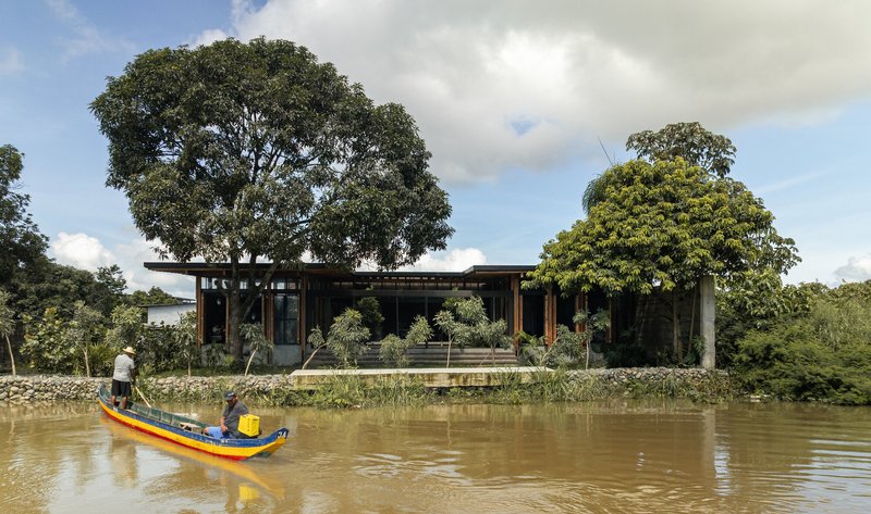 Riverside facade with wide overhanging roof and timber columns viewed from a canoe in muddy water