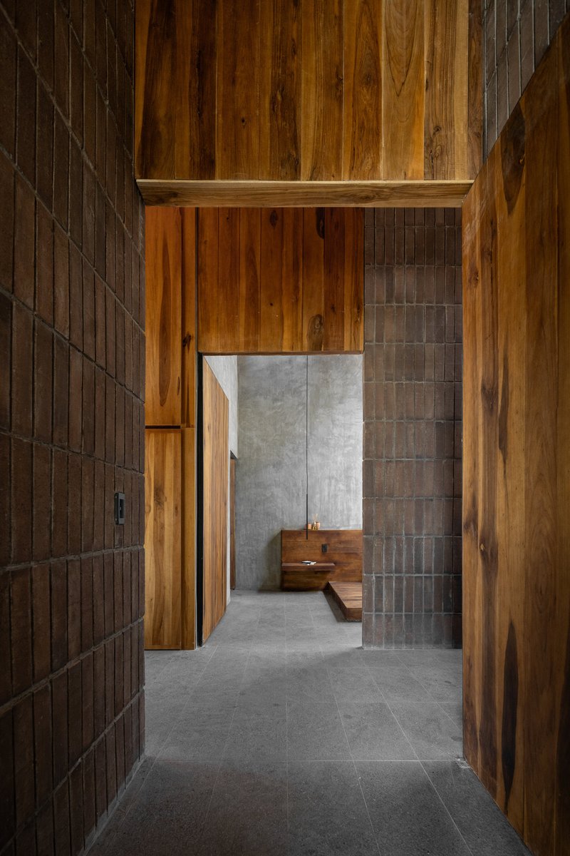 Corridor with vertical timber cladding and dark brick walls leading to a concrete-paneled room