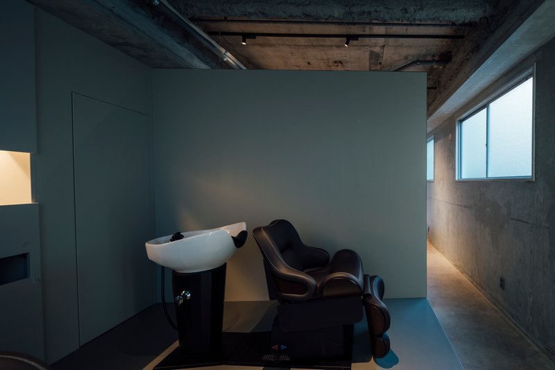 Salon space with wall-mounted washbasin, reclining chair, and concrete surfaces lit by natural light