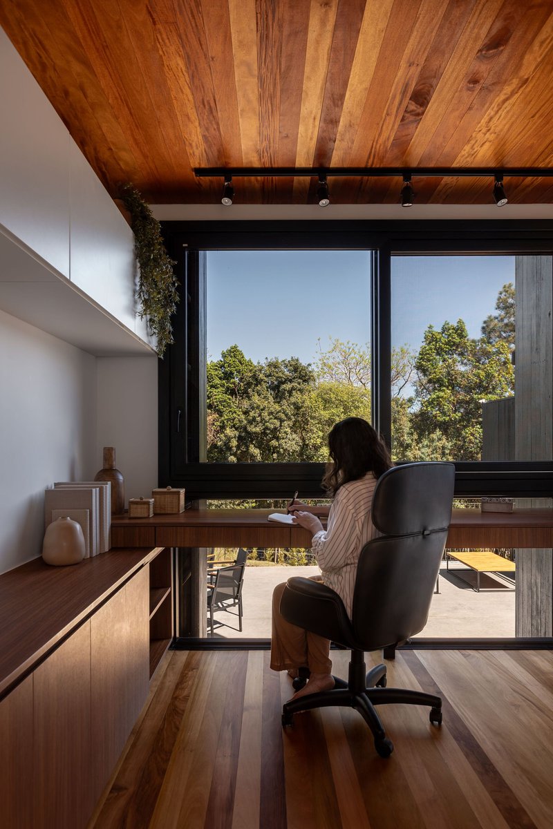 Home office with timber desk facing floor-to-ceiling windows overlooking green treetops in daylight