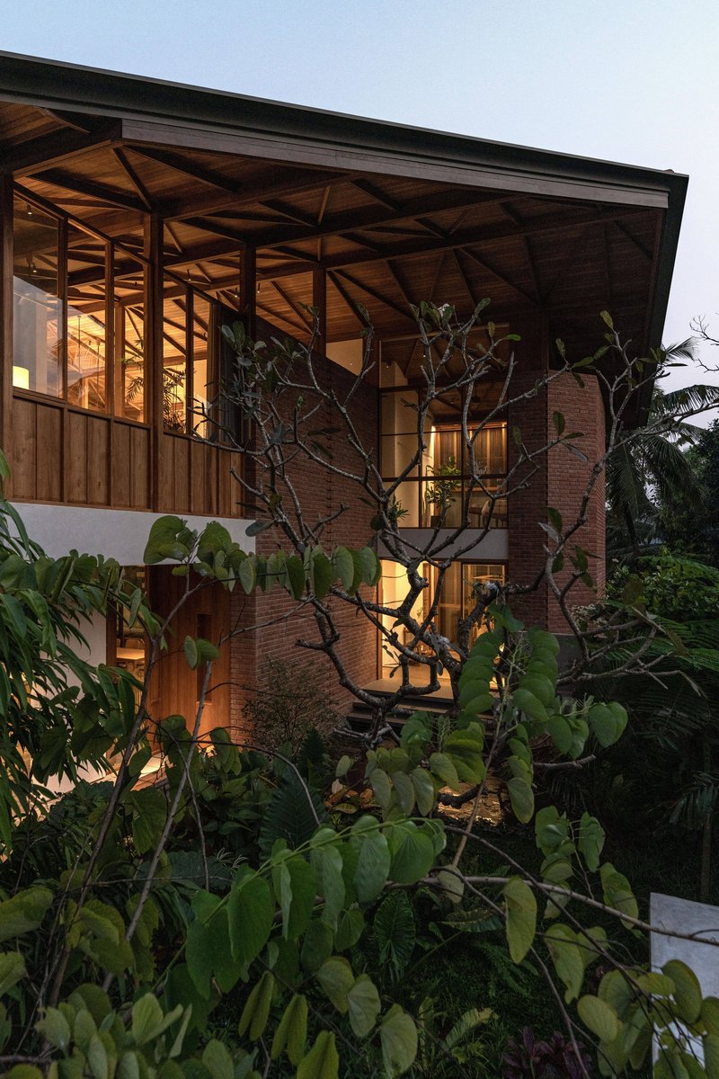 Evening view of the illuminated timber frame structure rising through planted garden with frangipani tree