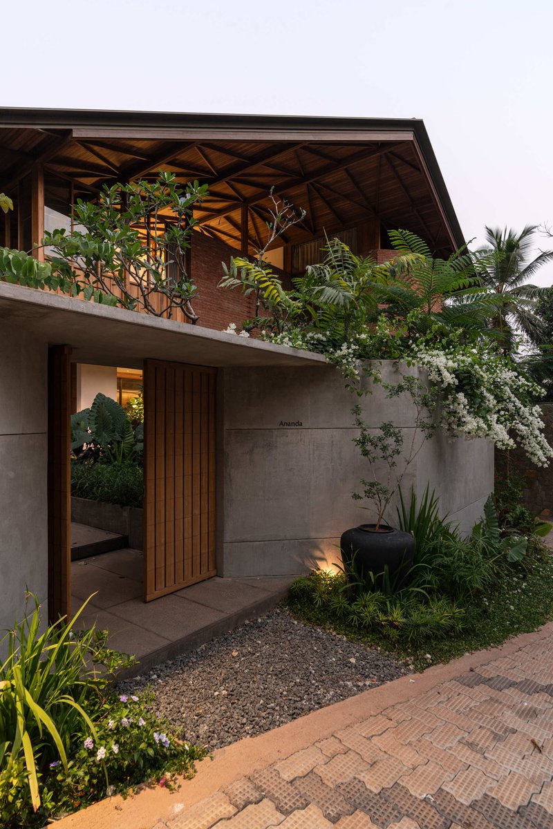 Entry facade with timber slat door and concrete walls beneath an upper terrace planted with tropical vegetation
