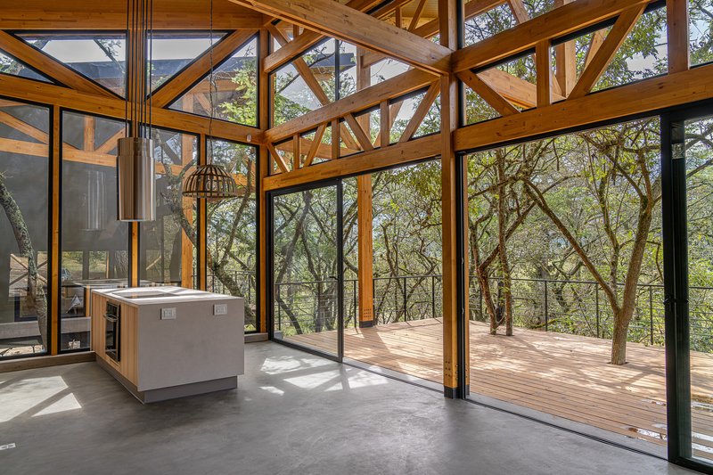 Kitchen with concrete island opening through floor-to-ceiling glazing onto a timber deck surrounded by trees