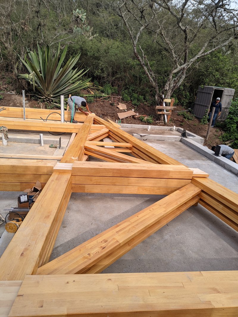 Close-up of timber roof trusses laid across concrete slab with worker visible in wooded background