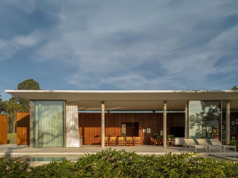 Covered poolside terrace with slender columns, timber-clad wall and recessed glass doors under afternoon light