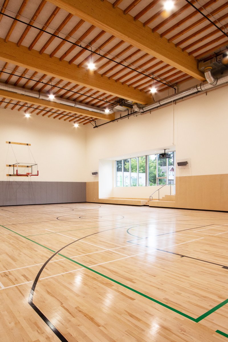 Basketball court with exposed timber beams, ductwork, and a wall-mounted hoop beneath clerestory windows