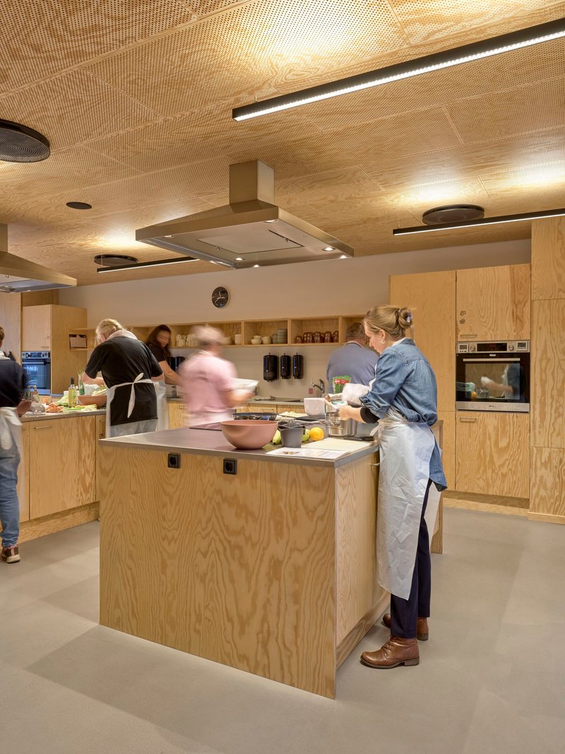 Communal kitchen with plywood island and cabinetry as people prepare food beneath a perforated timber ceiling