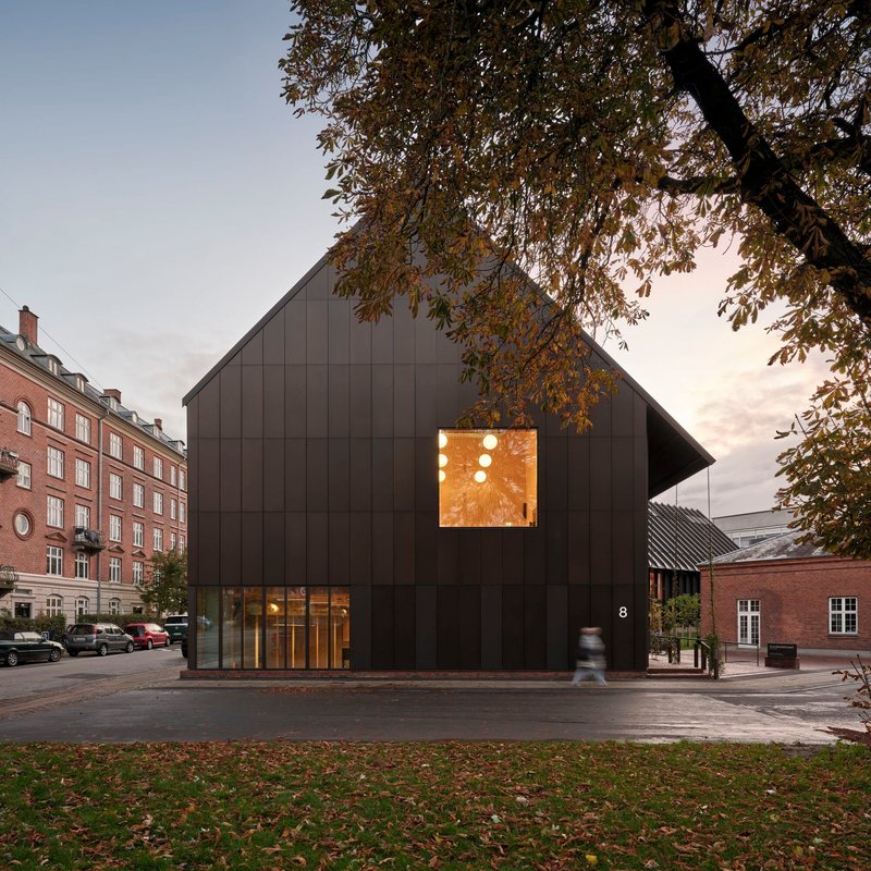 Dark metal-clad gabled facade with illuminated upper window and ground-level glazing at dusk