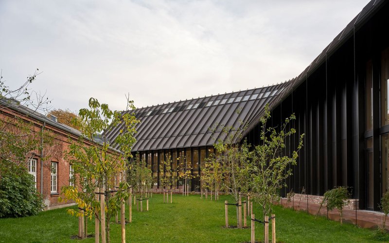 Sloped metal roof connecting to brick outbuilding with young trees planted in lawn