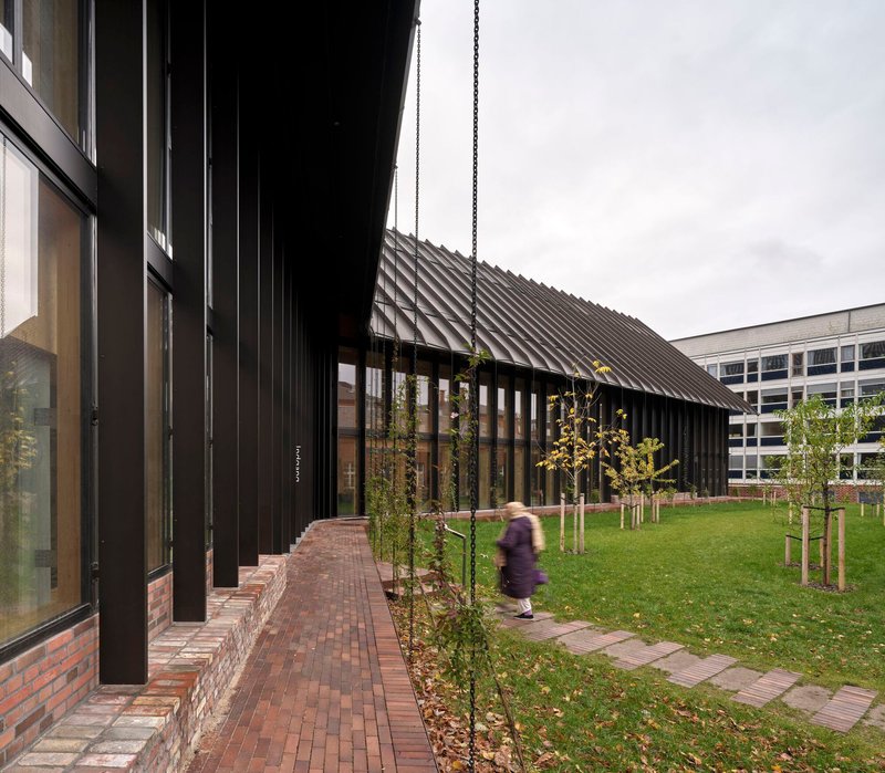 Brick walkway along black metal facade with young trees and a person passing by