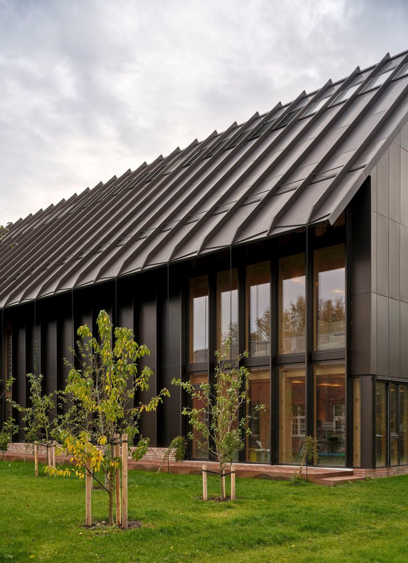 Sawtooth metal roof above glazed facade and black vertical fins with young trees on lawn in autumn