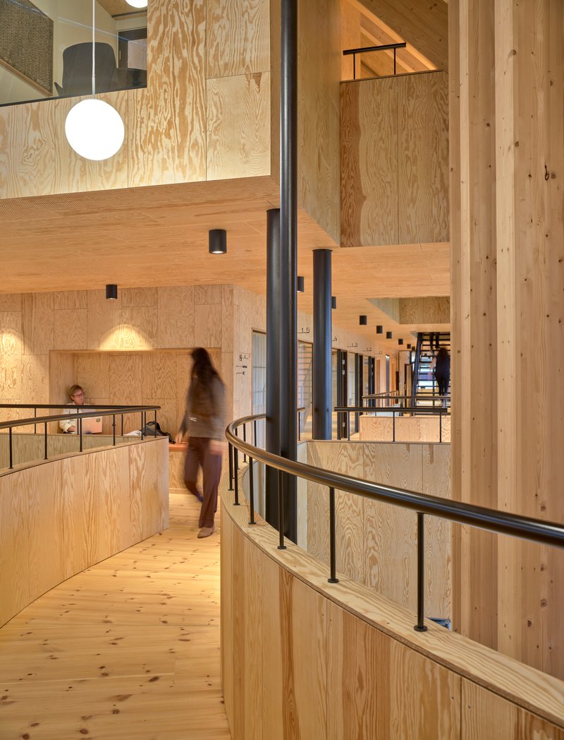 Curved balcony with black steel railings and a person in motion crossing the plywood-lined circulation level