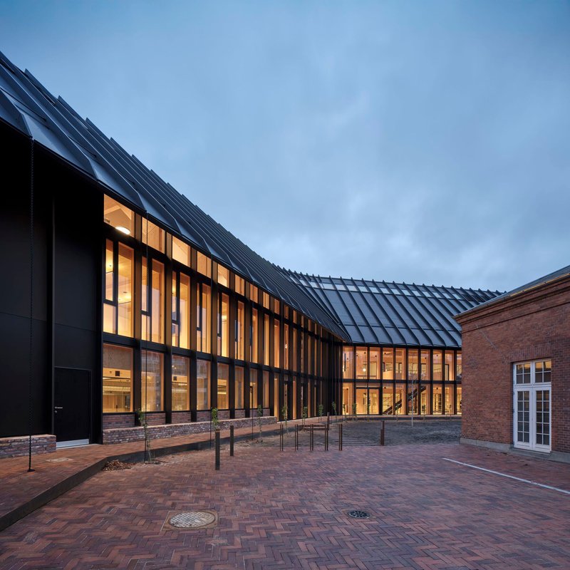 Brick plaza at dusk framed by the angular metal roof and warmly lit interiors
