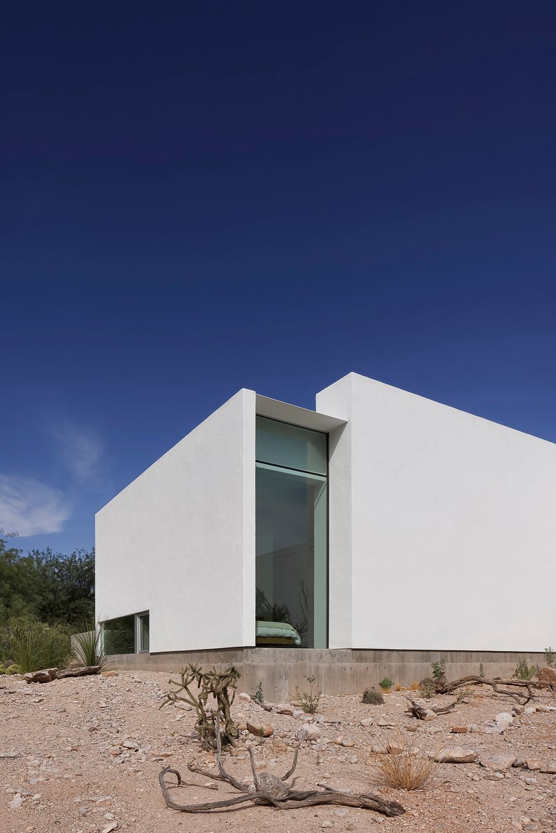 White stucco volumes framing a tall glazed slot window above desert terrain with scattered cacti