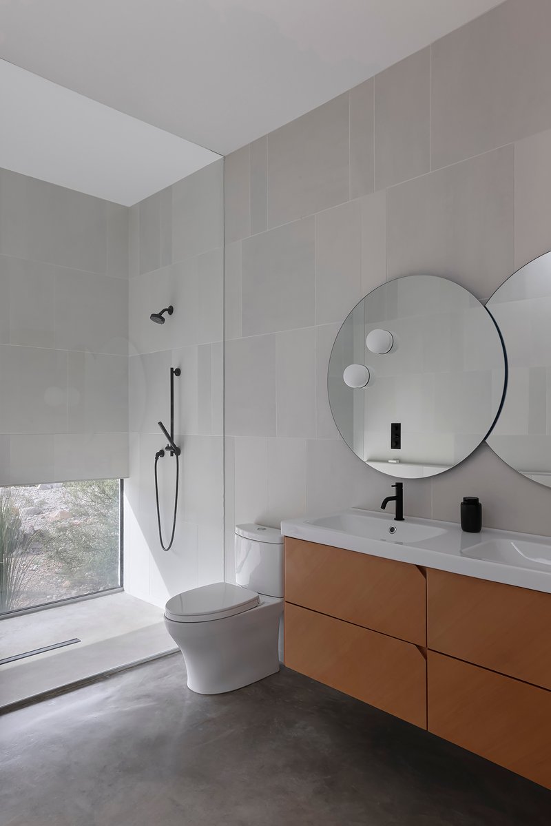 Bathroom with circular mirror above timber vanity and low desert window with roller shade