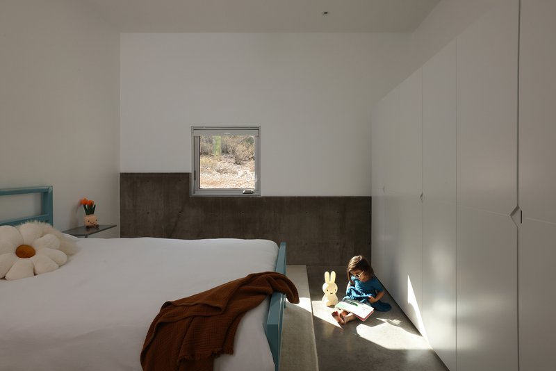 Child seated in sunlit corner beside bed with metal wainscoting and square desert window