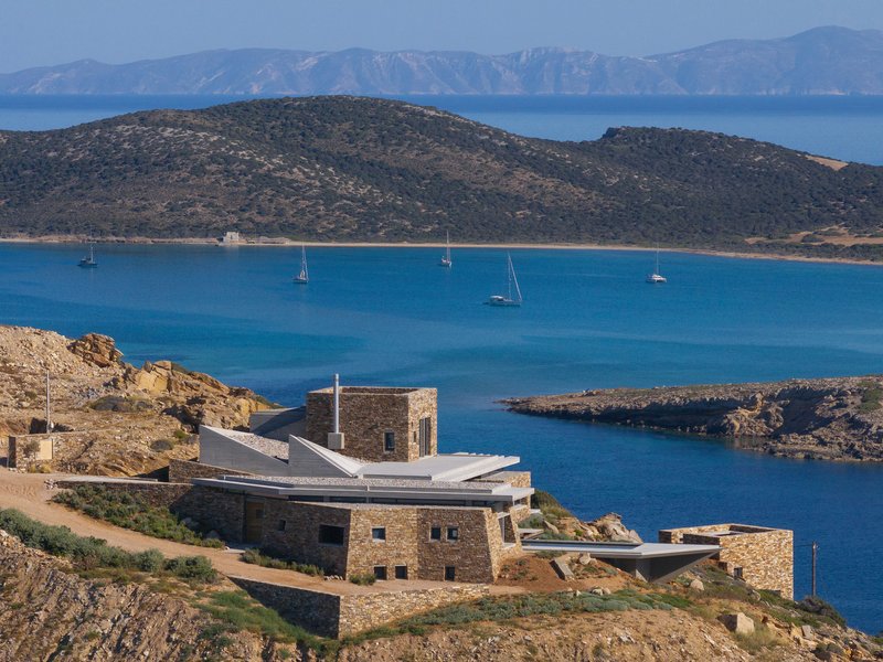 Hillside compound with stone walls and folded roof planes framing a bay with anchored sailboats