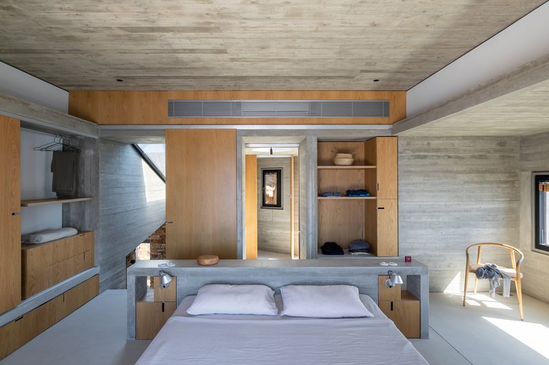 Bedroom with board-formed concrete walls, timber built-in wardrobes, and a doorway framing the passage beyond